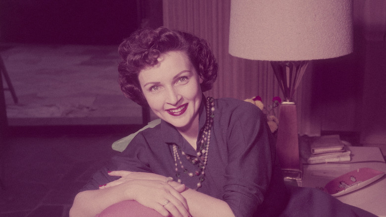 Betty White, actriz y comediante estadounidense, sonriendo mientras está sentada con los brazos cruzados sobre una almohada en el respaldo de un sofá en una sala de estar, alrededor de 1955.