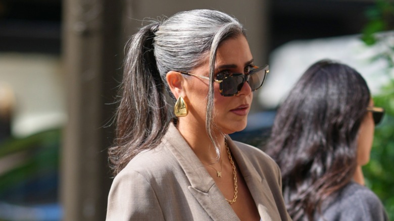 Una mujer con cabello gris recogido en una coleta alta, con gafas de sol, joyas de oro y una chaqueta gris.