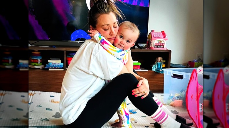 Kaley Cuoco con calzas y una camiseta abrazando a su hija pequeña Matilda, sentada en una alfombra con juguetes a su alrededor.