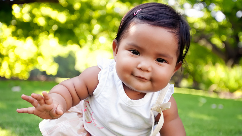 Niña sonriente con un traje blanco y un pasador rosa arrastrándose sobre la hierba.