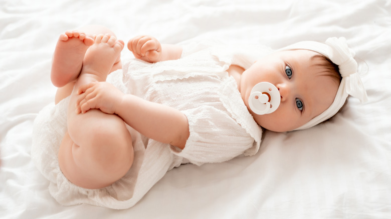 Niña con un chupete vestida de blanco sobre una manta blanca.