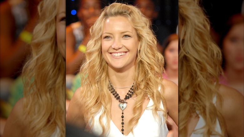 Kate Hudson con un chaleco corto blanco con un collar grueso negro y el cabello suelto con ondas sueltas con un puf.
