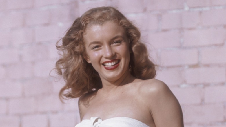 Marilyn Monroe posando con un top blanco, lápiz labial rojo y cabello rubio fresa ondulado frente a una pared de ladrillos.