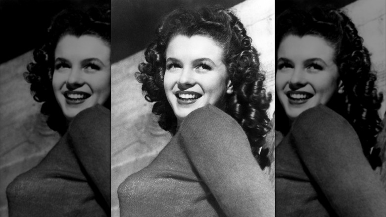 Fotografía en blanco y negro de Marilyn Monroe sonriendo y mirando por encima del hombro con cabello castaño y rizado.