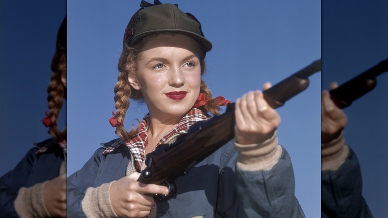 Marilyn Monroe posando en 1946 sosteniendo un rifle, vistiendo una camisa a cuadros, un sombrero de caza y lápiz labial rojo con el cabello recogido en trenzas.