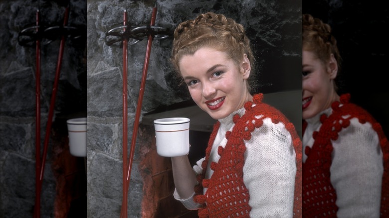 Marilyn Monroe posando en 1946 sosteniendo una taza con el cabello recogido en una trenza y vistiendo un chaleco de punto rojo sobre un suéter blanco.