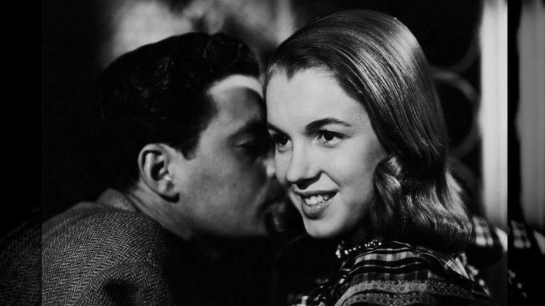Fotografía en blanco y negro de Marilyn Monroe con cabello castaño y liso sonriendo mientras un hombre le susurra al oído.