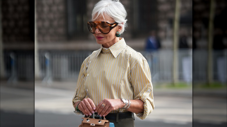 Una mujer mayor vestida con una mezcla de metales y un top amarillo y blanco con rayas.