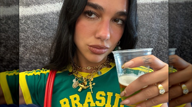 Selfie de Dua Lipa con un collar con dijes y un anillo llamativo mientras sostiene un vaso de cerveza