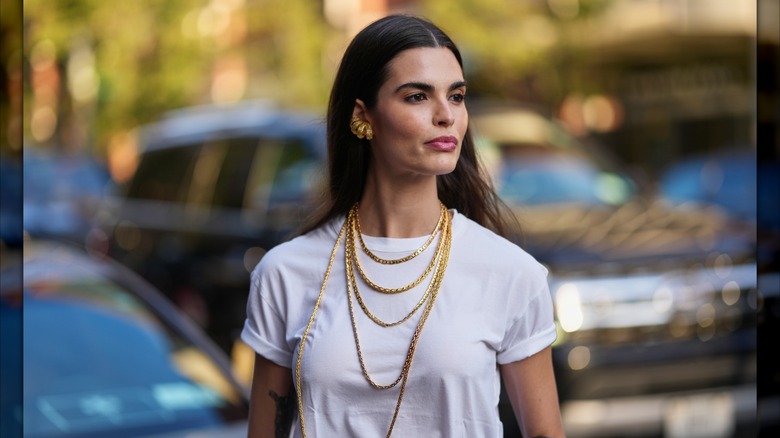 Una mujer que vestía una camiseta blanca con mangas remangadas y varios collares de cadenas doradas.