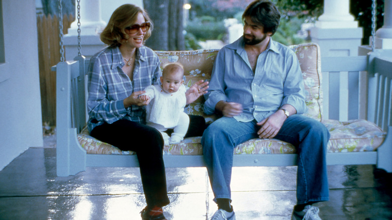 Cybill Shepherd en un columpio del porche con una camisa azul y sosteniendo a su bebé junto a David M. Ford.
