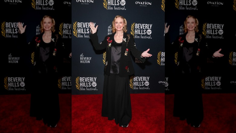 Cybill Shepherd sonríe con los brazos abiertos luciendo un blazer negro floral en un evento de alfombra roja.