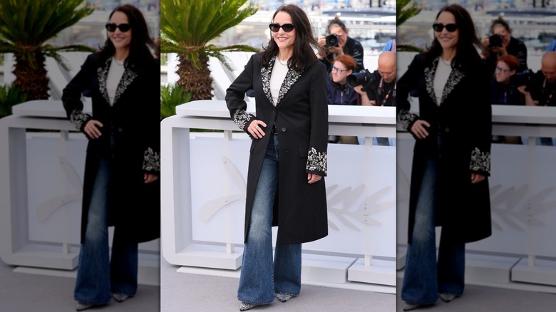 Julia Louis-Dreyfus posando en un evento con la mano en la cadera luciendo unos vaqueros de campana, un abrigo negro y gafas de sol oscuras.