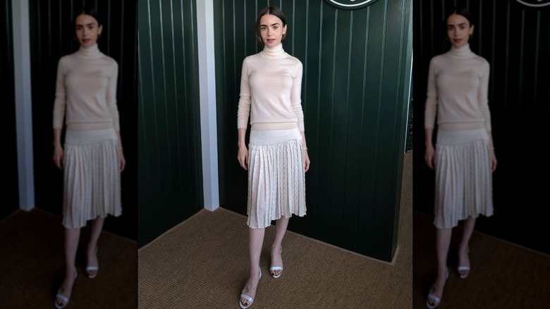 Lily Collins luciendo un top de cuello alto color crema y una falda plisada blanca con sandalias blancas en un evento.