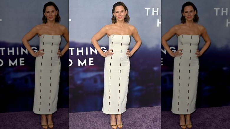 Jennifer Garner con los brazos en jarras luciendo un vestido palabra de honor blanco con diseños de guiones en un evento de alfombra roja.