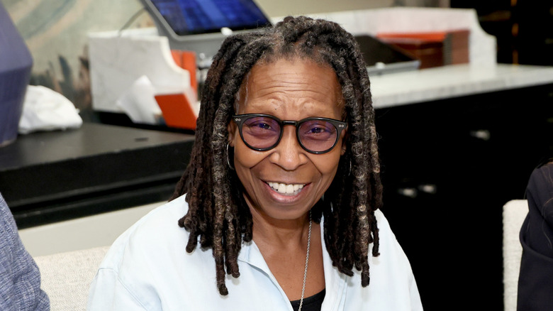Whoopi Goldberg sonriendo en un evento con una camisa blanca y lentes negros.