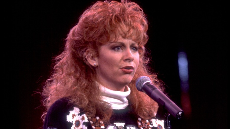 Reba McEntire canta frente a un micrófono mientras luce un voluminoso peinado rizado