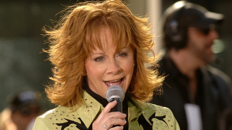 Reba McEntire canta y luce un corte de pelo con flecos en capas mientras usa una chaqueta de cuero verde claro.