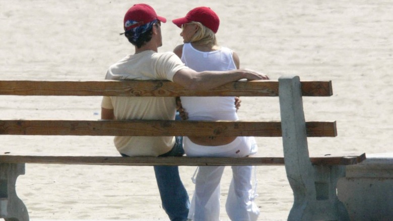 Anna Kournikova y Enrique Iglesias sentados en un banco de la playa con gorras rojas de béisbol