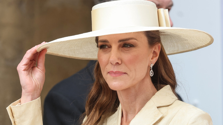 Kate Middleton tocándose el ala de su sombrero amarillo luciendo un blazer a juego con pendientes de diamantes.