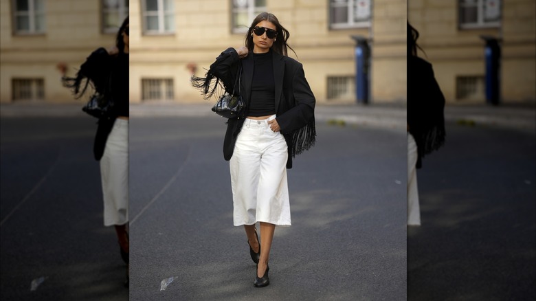 Una mujer vestida con una chaqueta negra con flecos y pantalones culottes blancos de pierna ancha.