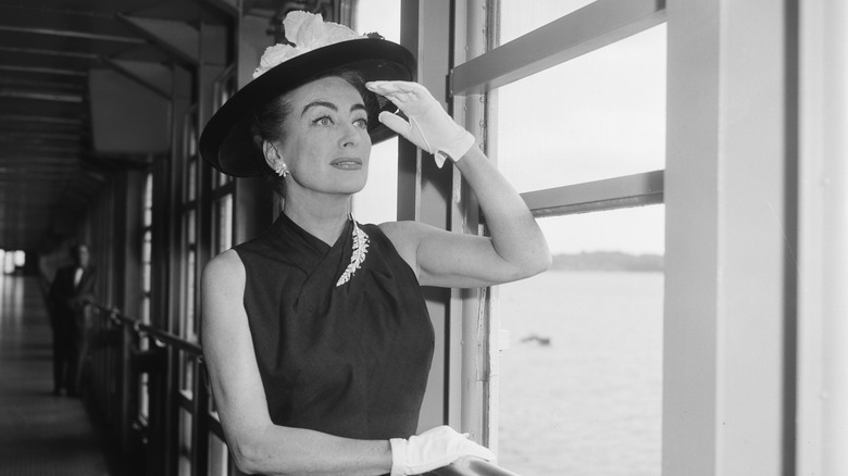 Joan Crawford mirando por la ventana de un barco con sombrero de ala ancha, vestido negro y guantes blancos.