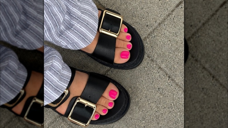 Primer plano de los pies de una mujer, con esmalte de uñas de color rosa intenso y sandalias negras gruesas
