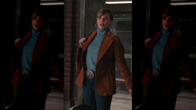 Belle Skinner con un jersey de cuello alto turquesa y un blazer de ante marrón