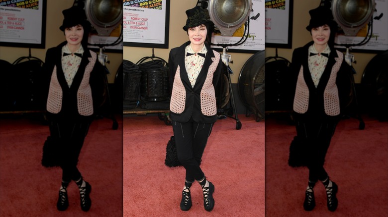 Toni Basil posando en la alfombra roja con los tobillos cruzados y luciendo un conjunto negro con detalles en rosa y blanco.