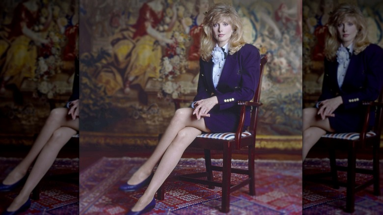 Morgan Fairchild sentada en una silla con las piernas cruzadas y vistiendo un traje de falda de color violeta oscuro.