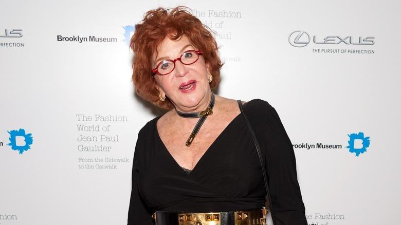 Sally Jessy Raphael llevaba un top negro escotado con un cinturón dorado brillante y un collar con cinturón a juego junto con su cabello corto rojo y sus característicos lentes rojos mientras asistía al Fashion World of Jean Paul Gaultier en la ciudad de Nueva York en 2013.