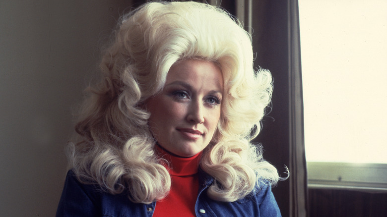 Dolly Parton con una voluminosa peluca rubia, un jersey de cuello alto rojo y una camisa azul.
