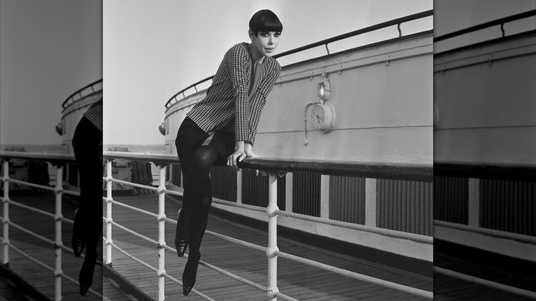 Peggy Moffitt posando en la barandilla de un barco luciendo una chaqueta de cuadros con falda negra y medias.