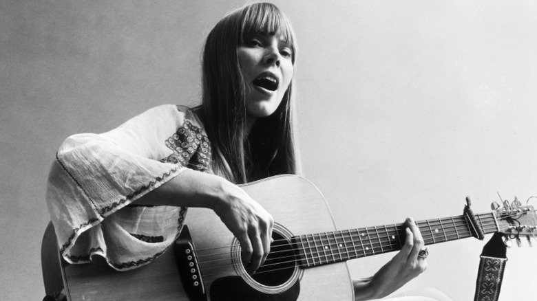 Un retrato de Joni Mitchell sentada en el suelo tocando la guitarra.