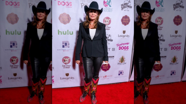 La actriz Kelly LeBrock en la alfombra roja vistiendo una camisa blanca con una chaqueta y pantalón negros con un sombrero de vaquero negro y botas de vaquero rojas.