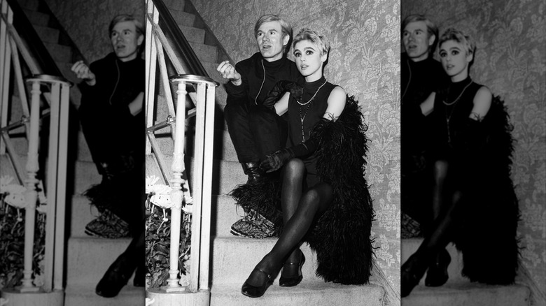 El artista Andy Warhol vestido completamente de negro y Edie Sedgwick vistiendo un traje negro con una boa de plumas negra, sentados en una escalera.