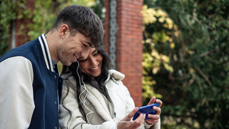 Una pareja usando audífonos y riéndose de la pantalla de un teléfono inteligente