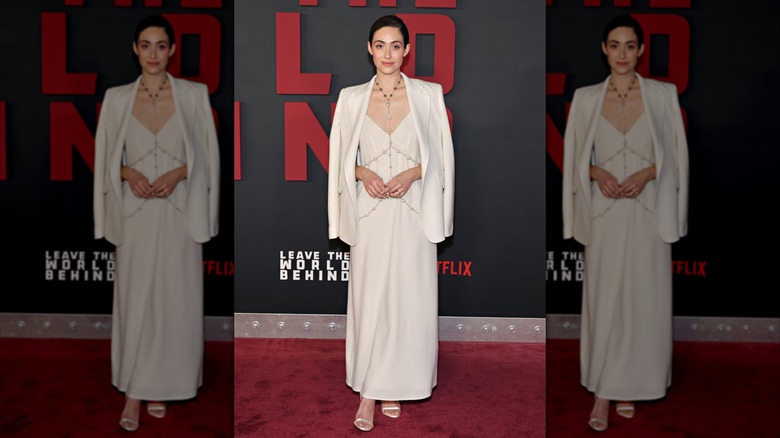Emmy Rossum lleva una chaqueta blanca y un vestido con una larga cadena plateada.