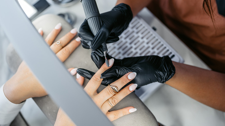 Mujer recibiendo sus uñas acrílicas de manicura francesa pulidas por un técnico con guantes negros en un salón.