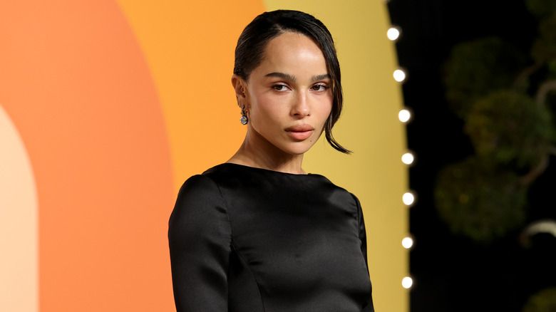Zoë Kravitz posa frente a un fondo naranja y amarillo mientras lleva un vestido negro y un recogido.
