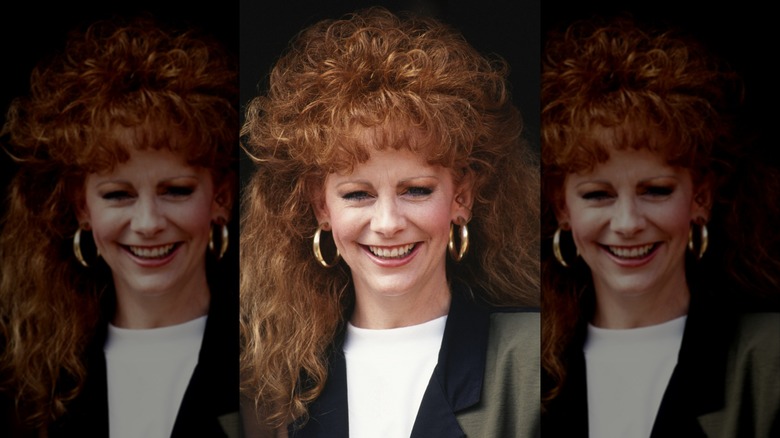 Reba McEntire con el pelo largo y rizado de color rojo y vistiendo un top blanco y una chaqueta en 1990.