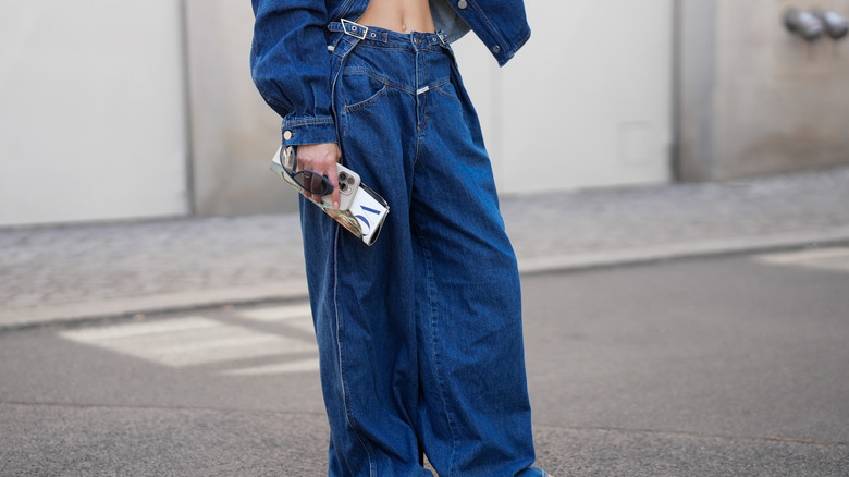 Una mujer usa jeans holgados de talle bajo y chanclas mientras sostiene una revista Vogue, su teléfono y sus gafas de sol.