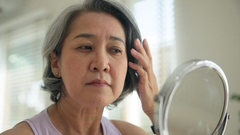Mujer madura con cabello canoso mira en un pequeño espejo redondo