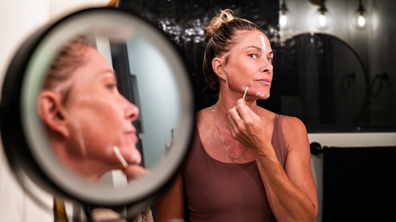 Una mujer de mediana edad con el pelo recogido en un moño se aplica crema para el cuidado de la piel en la cara con un hisopo de algodón
