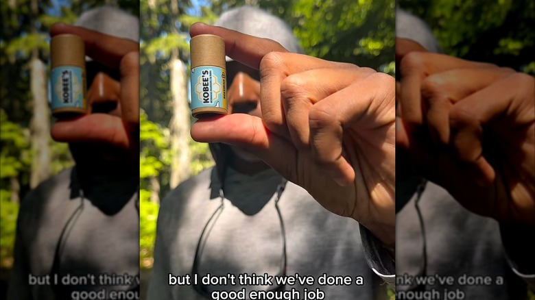 Kobe Harris con una sudadera con capucha gris sosteniendo uno de los bálsamos labiales de su marca.