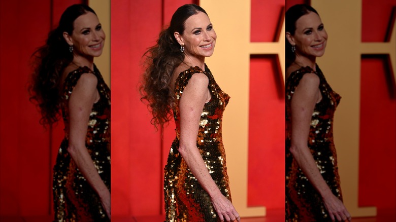 Minnie Driver lleva un vestido dorado brillante con el pelo recogido en una cola de caballo peinada hacia atrás.