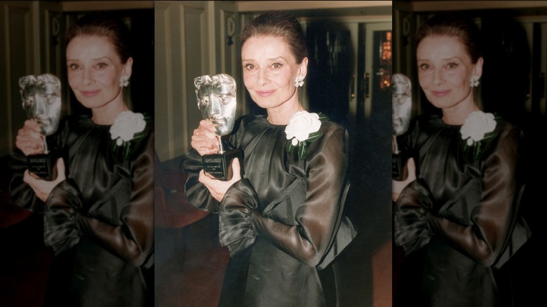 Audrey Hepburn posando con un premio con un vestido negro con mangas transparentes y un detalle de rosa blanca.