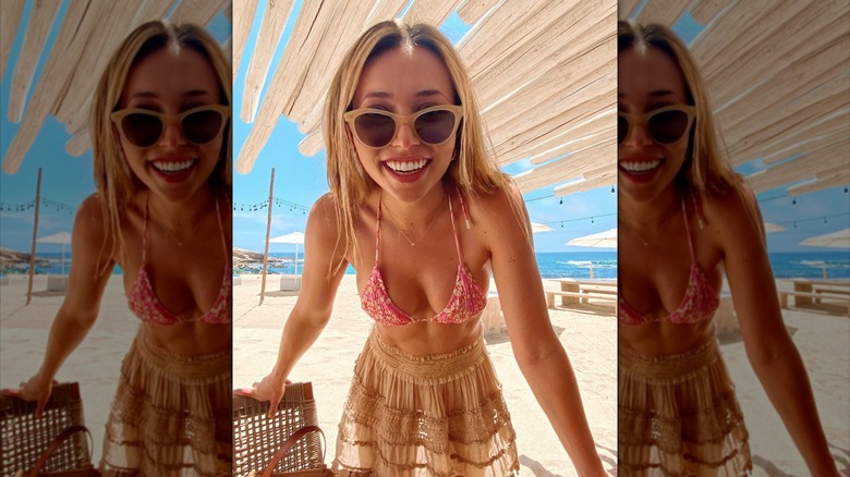 Camelia Somers usa gafas de sol, un top de bikini floral rosa y una falda color canela transparente en la playa.