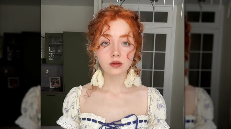 Una pelirroja con maquillaje cottagecore y aretes de flores posando para una foto.