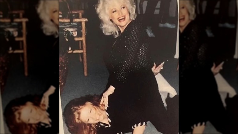 Judy Ogle yace debajo de Dolly Parton vistiendo un conjunto negro y su cabello pelirrojo extendido por el suelo.